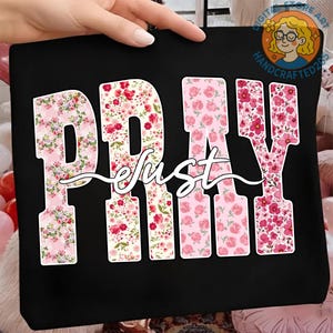 May include: Black tote bag with the word "PRAY" in large, floral-patterned letters. The word "just" is written in cursive script across the center. The floral patterns include pink and red flowers on a white background.