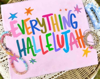 May include: A light pink t-shirt featuring the colorful text EVERYTHING HALLELUJAH in a playful font. The design is accented with small, multi-colored stars in blue, orange, pink, and green. A beaded necklace and a blue checkered hair clip are present.