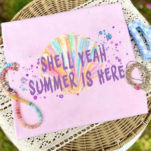 May include: A light purple t-shirt featuring a central graphic of a multi-colored iridescent seashell. The text "SHELL YEAH SUMMER IS HERE" is printed in purple letters over the shell. Scattered purple and blue glitter accents surround the design.