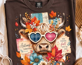 May include: A dark brown t-shirt featuring a graphic of a Highland cow wearing heart-shaped glasses with plaid lenses. The design includes colorful autumn leaves, flowers, mushrooms, and the text "Autumn leaves" and "the leaves fall one by one."