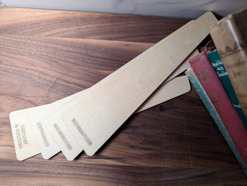 May include: Wooden bookmarks with engraved text, including "History & Culture," "Reference," "Workbooks," and "Biographies." The bookmarks are light-colored and arranged on a dark wooden surface, next to a stack of vintage books.