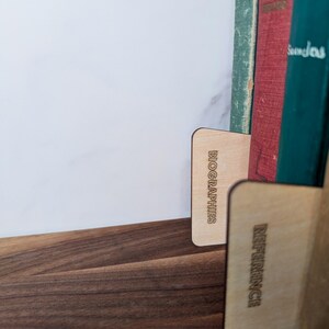 May include: Wooden bookends with the words "BIOGRAPHIES" and "REFERENCE" etched into them. The bookends are holding up a collection of vintage books with green, red, and blue covers. The bookends are on a dark wooden surface.
