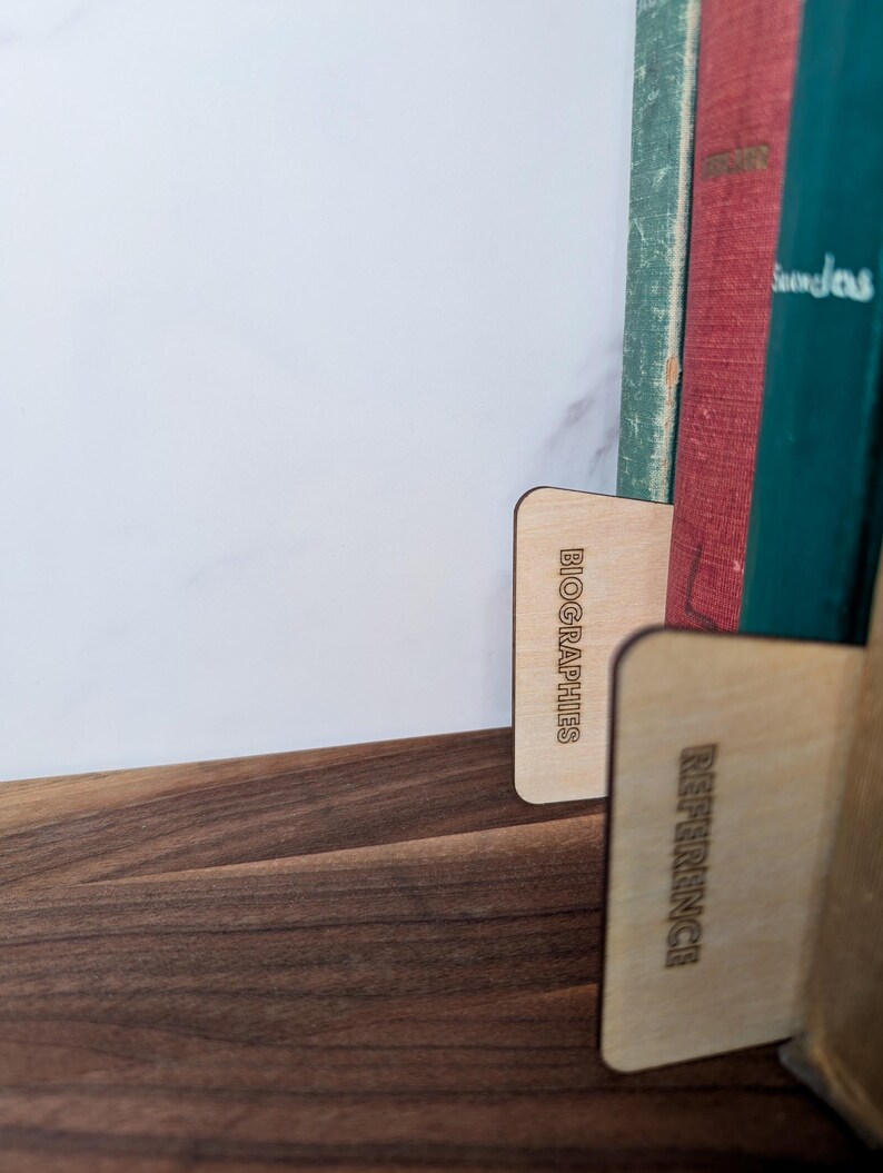 May include: Wooden bookends with the words "BIOGRAPHIES" and "REFERENCE" etched into them. The bookends are holding up a collection of vintage books with green, red, and blue covers. The bookends are on a dark wooden surface.