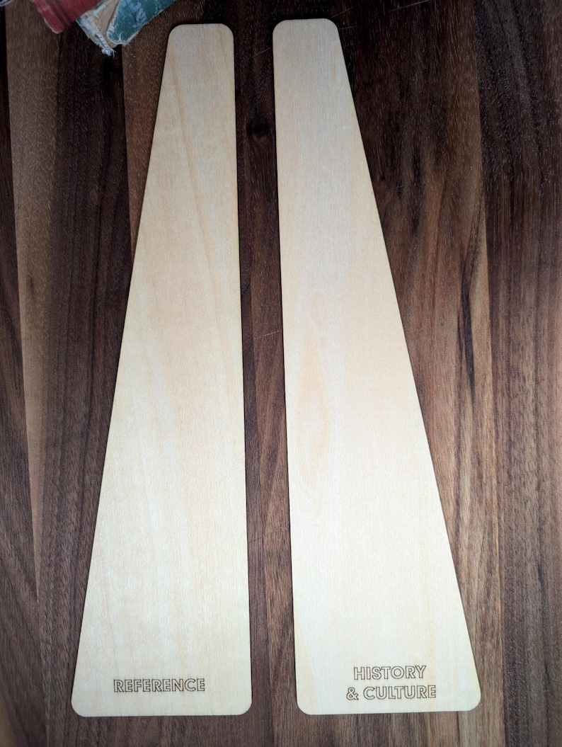 May include: Two light-colored wooden display stands with a trapezoid shape. One stand has the word "REFERENCE" etched into the wood, and the other has "HISTORY & CULTURE". The stands are on a dark wood surface.