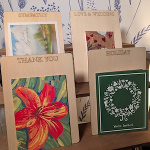 May include: Wooden card holders with engraved text for various occasions. The cards include a landscape scene, a heart-themed design, a red flower painting, and a green card with a wreath and the text "You're Invited".