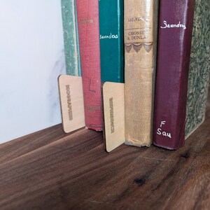 May include: A collection of vintage books with colorful spines, held in place by two wooden bookends. The books are arranged on a dark wooden shelf, creating a warm and inviting aesthetic. The bookends have text on them.