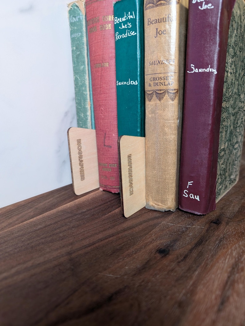 May include: A collection of vintage books with colorful spines, held in place by two wooden bookends. The books are arranged on a dark wooden shelf, creating a warm and inviting aesthetic. The bookends have text on them.