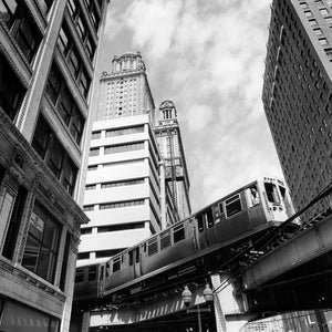 May include: Black and white photograph of a city scene featuring a train on elevated tracks. Tall buildings with ornate architectural details frame the train. The sky is overcast.