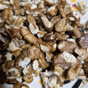 May include: Close-up of shelled walnuts, showcasing their light brown and beige hues. The nuts are arranged on a decorative plate with a patterned rim. The image highlights the texture and natural colors of the walnuts.