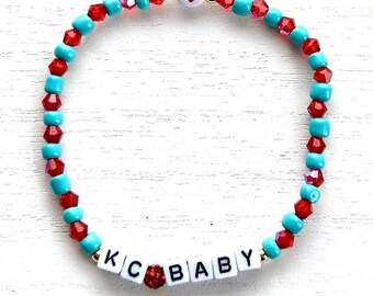 May include: A turquoise and red beaded bracelet with a gold heart charm and white letter beads spelling "KC BABY".
