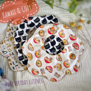 May include: Three flower-shaped baby bibs with snap closures. One bib features a ramen and Shiba Inu dog print on a cream background. Another bib displays a black cat pattern on a white background. The third bib shows a multi-colored cat print.