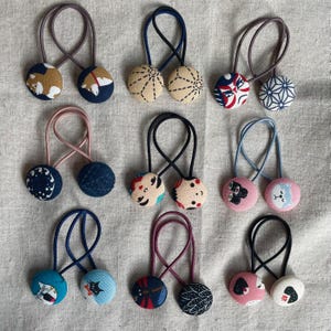 May include: A collection of handmade hair ties featuring fabric-covered buttons with various patterns and designs. The buttons are attached to elastic bands in different colors, including blue, pink, and black. The designs include geometric patterns, floral prints, and animal illustrations.