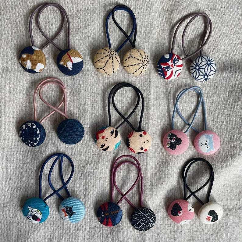 May include: A collection of handmade hair ties featuring fabric-covered buttons with various patterns and designs. The buttons are attached to elastic bands in different colors, including blue, pink, and black. The designs include geometric patterns, floral prints, and animal illustrations.
