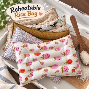 May include: A collection of rectangular fabric pouches labelled Reheatable Rice Bag, featuring various patterns including pink and red strawberries on white, blue floral, mustard yellow with white dots, and a black and white cat print.