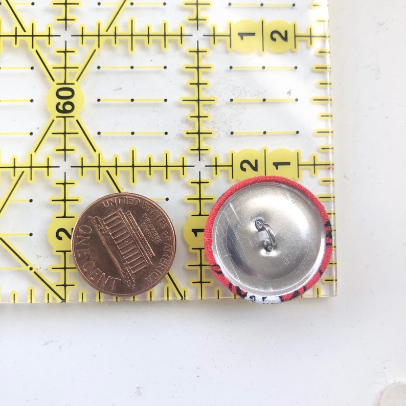 May include: A red and white fabric-covered button with a metal back, next to a penny and a yellow ruler. The button has a black and white pattern around the edge. The ruler is marked in inches.