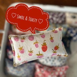 Könnte beinhalten: Ein rechteckiges Kissen mit einem Muster aus rosa und roten Erdbeeren mit grünen Stielen auf weißem Grund. Das Kissen hat einen gelben Rand. Eine rote Sprechblase mit dem Text "Smol & Toasty" befindet sich in der oberen linken Ecke.