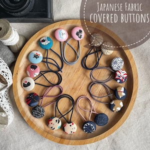 May include: A collection of Japanese fabric-covered buttons arranged on a wooden tray. The buttons feature various patterns and colors, including floral, geometric, and animal designs. Each button is attached to a hair tie. The text "Japanese Fabric Covered Buttons" is visible.