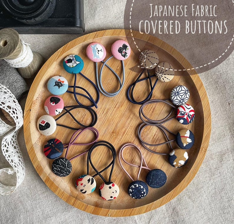 May include: A collection of Japanese fabric-covered buttons arranged on a wooden tray. The buttons feature various patterns and colors, including floral, geometric, and animal designs. Each button is attached to a hair tie. The text "Japanese Fabric Covered Buttons" is visible.