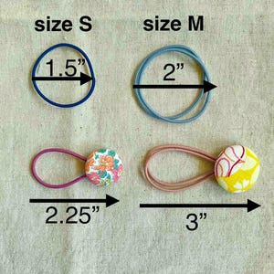 May include: An assortment of hair ties in various sizes and styles. Includes a blue elastic band measuring 1.5 inches, a light blue band at 2 inches, and two button-adorned ties at 2.25 and 3 inches. The buttons feature floral and abstract patterns.