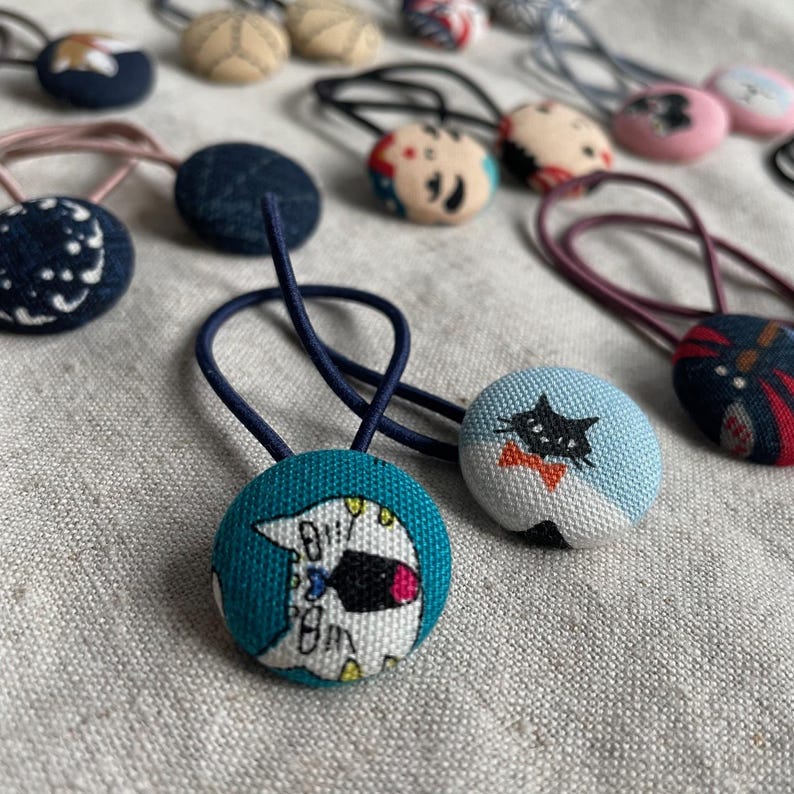 May include: A collection of fabric-covered hair ties with various patterns and designs. The ties feature round, button-like embellishments with images of cats, floral patterns, and abstract designs. The color palette includes blues, pinks, and neutral tones.