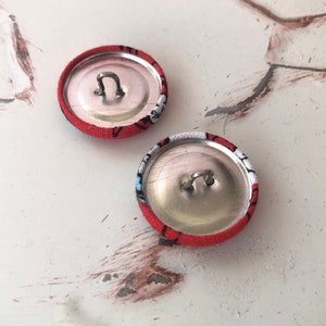 May include: Two fabric-covered buttons with a red background and a black and white pattern. The buttons have a silver metal loop on the back for sewing. The buttons are round and appear to be handmade.