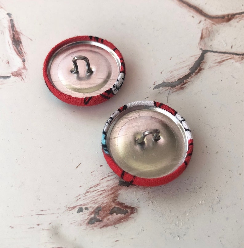 May include: Two fabric-covered buttons with a red background and a black and white pattern. The buttons have a silver metal loop on the back for sewing. The buttons are round and appear to be handmade.