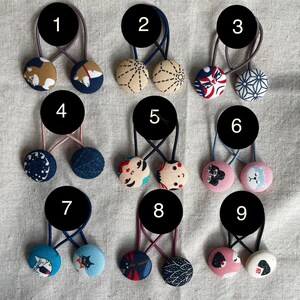May include: A collection of nine pairs of fabric-covered button hair ties. Each pair features two round buttons with various patterns and colors, including floral, geometric, and animal designs. The elastic bands are in various colors.