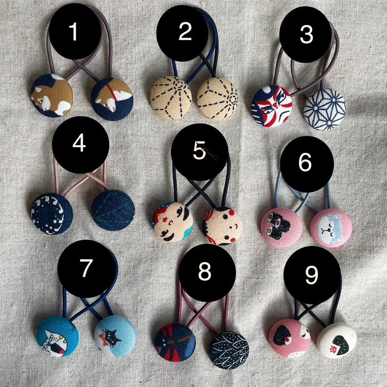 May include: A collection of nine pairs of fabric-covered button hair ties. Each pair features two round buttons with various patterns and colors, including floral, geometric, and animal designs. The elastic bands are in various colors.