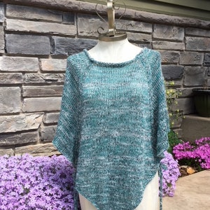 May include: A teal and white knitted poncho with a triangular shape and a fringe trim. The poncho is displayed on a mannequin against a stone wall background.