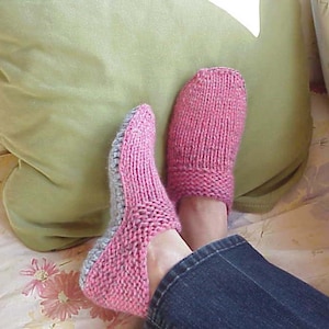 Puede incluir: Un par de zapatillas de punto rosas y grises. Las zapatillas son de color rosa suave con un ribete gris alrededor del talón y la puntera. Las zapatillas están en los pies de una persona, con vaqueros azules visibles.