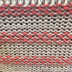 May include: Close-up of a knitting loom with white and pink yarn creating a striped pattern.