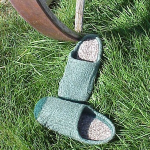 May include: A pair of green knitted slippers with a brown suede insole. The slippers are sitting on a bed of green grass.