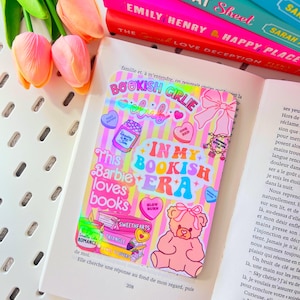 Op de afbeelding: Een kleurrijke sticker met de tekst "BOOKISH GIRLIE CLUB" en "IN MY BOOKISH ERA" wordt weergegeven op een open boek. De sticker bevat een roze teddybeer, harten en andere afbeeldingen. Een stapel boeken en roze tulpen zijn op de achtergrond.