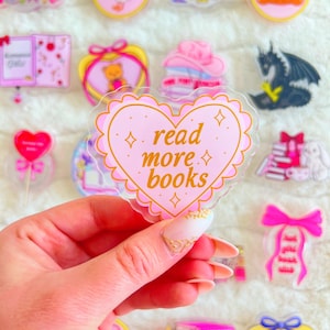 May include: A collection of pink and clear acrylic pins with various designs, including a heart-shaped pin with the text "read more books". Other pins feature a teddy bear, a goose, a dragon, and a stack of books. The pins are on a white, fluffy surface.