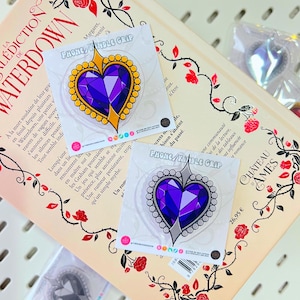 Pode incluir: Suportes para telefone/Kindle em forma de coração com um design de gema roxa, rodeados por acabamentos dourados ou prateados. Os suportes estão em um livro com detalhes florais e o texto "WATERDOWN".