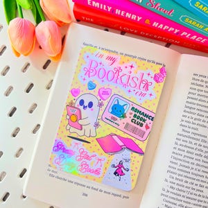 Peut inclure: Un livre ouvert avec un marque-page coloré indiquant « in my Bookish era ». Le marque-page présente des illustrations mignonnes et le texte « Romance Book Club ». Des tulipes roses et une pile de livres sont en arrière-plan.