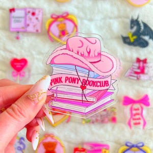 May include: A collection of pink and clear acrylic charms and stickers. The central charm features a pink cowboy hat atop a stack of books with the text "PINK PONY BOOKCLUB". Other charms include a teddy bear, a goose, and a heart.