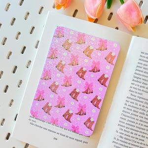 Può includere: Un adesivo rettangolare rosa con un motivo di stivali da cowboy marroni con fiocchi rosa e accenti di margherite bianche. L'adesivo è su un libro aperto con testo, con tulipani rosa sullo sfondo.