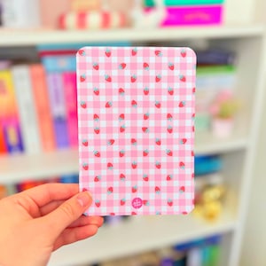 Könnte beinhalten: Ein rechteckiger Notizblock mit abgerundeten Ecken, der ein rosa-weißes Vichy-Muster aufweist. Kleine rote Erdbeeren mit grünen Blättern sind über das Design verstreut. Der Notizblock wird in einer Hand gehalten, mit einem verschwommenen Bücherregal im Hintergrund.