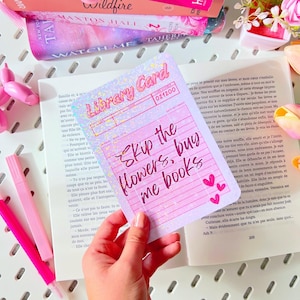 Op de afbeelding: Een roze bibliotheekkaart met de tekst "Skip the flowers, buy me books" wordt boven een open boek gehouden. De kaart heeft een holografisch effect en een bibliotheeketiketontwerp. Roze boeken en markers staan op de achtergrond.