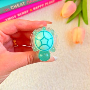 Peut inclure: Un support de téléphone en forme de tortue turquoise et bleu sarcelle. La poignée a une base circulaire transparente et est tenue dans une main. En arrière-plan, des livres et des tulipes roses et pêche.