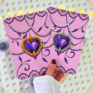 Pode incluir: Dois cartões rosa em forma de coração com desenhos ornamentados. Cada cartão apresenta uma grande joia roxa em forma de coração com detalhes dourados ou prateados, rodeada por elementos decorativos. Os cartões são segurados em uma mão.