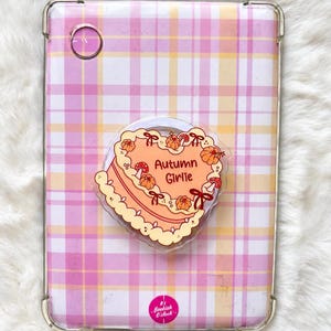 Peut inclure: Une coque de tablette à motif écossais rose, blanc et jaune avec un support de téléphone en forme de cœur. Le support présente une illustration de gâteau pêche avec le texte Autumn Girlie, orné de petites citrouilles, champignons et nœuds rouges.