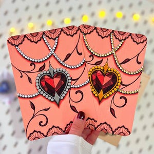 Pode incluir: Dois porta-cartões corais com desenhos de pedras preciosas em forma de coração. Um coração é prateado, o outro dourado. Ambos têm pedras vermelhas, detalhes de pérolas e padrões florais pretos. Os porta-cartões são segurados por uma pessoa.