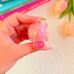 Pode incluir: Um tampão de garrafa rosa em forma de tartaruga. O tampão é segurado em frente a um fundo de livros e flores. A tartaruga tem um corpo transparente e redondo com um design de casco de tartaruga rosa e uma cabeça redonda rosa.