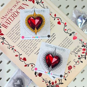 Pode incluir: Punhos de telefone/alça em forma de coração com um design de gema vermelha, acentuados com detalhes dourados e prateados. Os punhos são exibidos em um livro com ilustrações florais e o texto "PHONE/HANDLE GRIP".