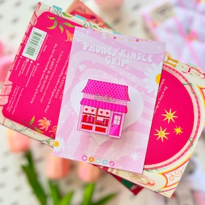 Peut inclure: Un support de téléphone/Kindle sur le thème d'une librairie rose et rouge. Le support représente un petit bâtiment avec un toit rose et le mot "Bookstore" sur le devant. Le support est sur un fond rose avec les mots "PHONE/KINDLE GRIP".