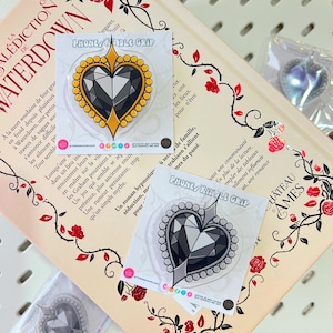 Pode incluir: Suportes de telefone em forma de coração com design de gema. Um tem borda dourada, o outro prateada. Os suportes estão em um livro com ilustrações florais e o texto "PHONE/KINDLE GRIP".