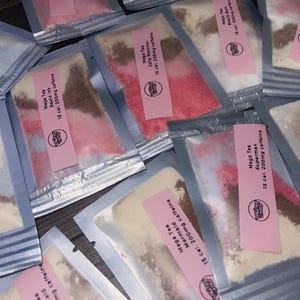 May include: An overhead shot of several sealed packets of Mega Tea. Each packet has a pink label with the brand logo and flavor name, such as "Mermaid" and "Jolly Rancher." The packets contain colorful powders, with text indicating 15 calories and 200mg of caffeine.
