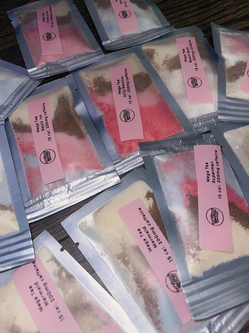 May include: An overhead shot of several sealed packets of Mega Tea. Each packet has a pink label with the brand logo and flavor name, such as "Mermaid" and "Jolly Rancher." The packets contain colorful powders, with text indicating 15 calories and 200mg of caffeine.